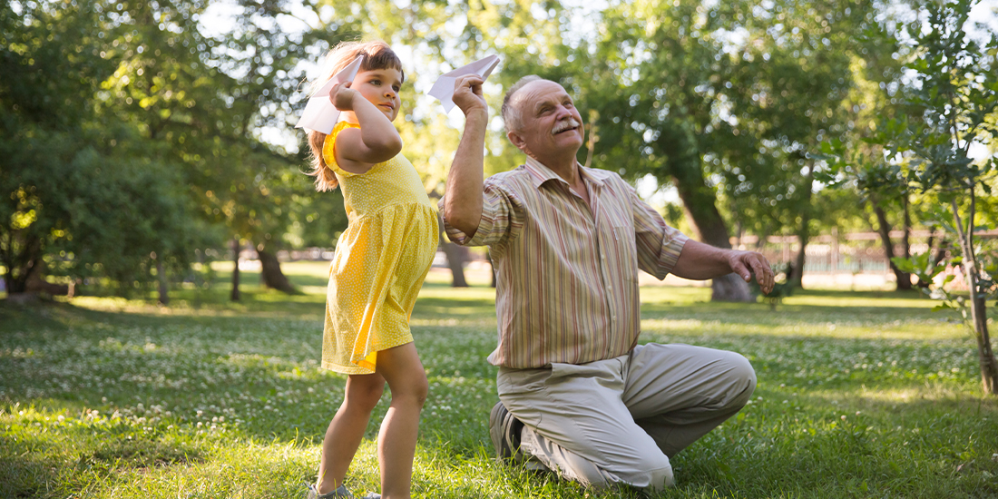 Ein Senior und ein kleines Mädchen im gelben Kleid spielen im Park mit Papierflugzeugen Ein Senior und ein kleines Mädchen im gelben Kleid spielen im Park mit Papierflugzeugen
